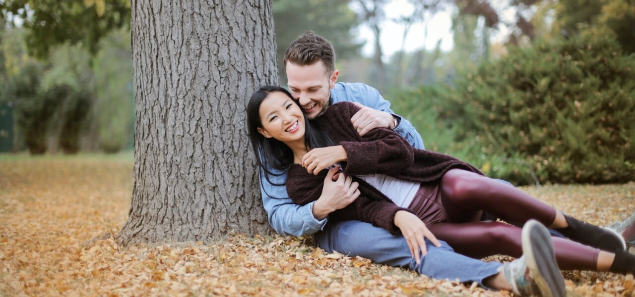 A happy couple sits in a park, embracing each other while sitting under a tree.