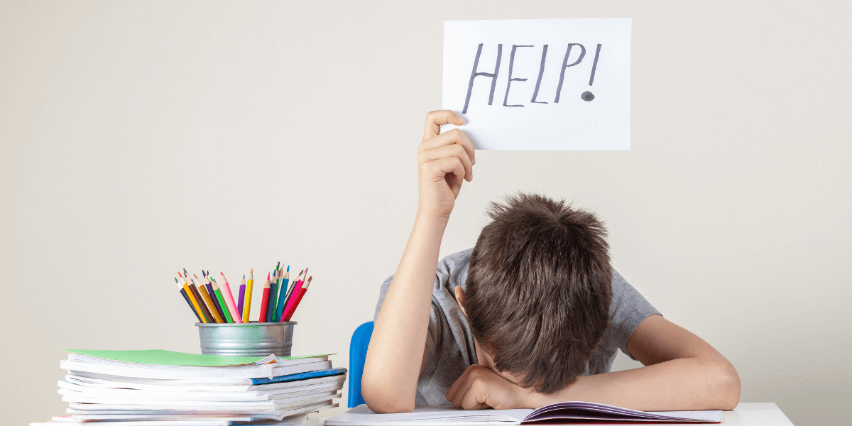 Kid laying his head down in a study desk holing a help sign with his left hand
