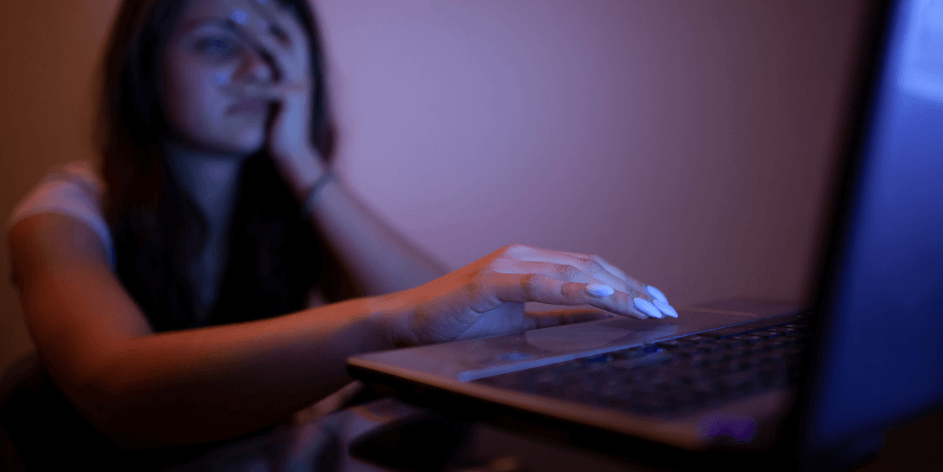 A woman using her laptop with one hand and touching her face with another hand in dark room.